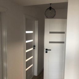 Solutionis Marek Skałbania - Białe drzwi z czarnymi klamkami i przeszkleniami w jasnym korytarzu z drewnianą podłogą. Nad drzwiami wisi czarna, druciana lampa.