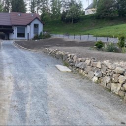 Stany surowe, żelbet, stropy monolityczne, domy i usługi. Śląsk Chorzów, Katowice okolice. - Nowo wykończony podjazd z kamienną ścianą oporową prowadzący do nowoczesnego domu z ciemnym dachem i dużymi oknami, otoczony zielenią i świeżo przygotowanym terenem.