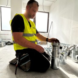 Stany surowe, żelbet, stropy monolityczne, domy i usługi. Śląsk Chorzów, Katowice okolice. - Instalator w żółtej kamizelce łączy elementy wentylacji w surowym wnętrzu. Widoczne liczne, metalowe rury i narzędzia. Jasne światło wpada przez okno.