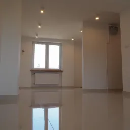 Puste, jasne wnętrze mieszkania po remoncie, z błyszczącą, beżową podłogą, białymi ścianami i sufitowym oświetleniem punktowym, widoczny grzejnik pod oknem i licznik gazu na ścianie.