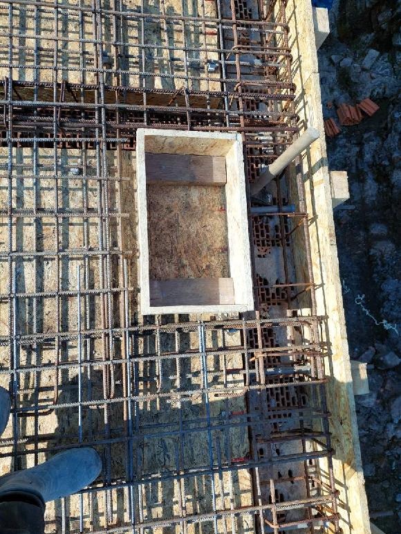 Zbrojenie stropu z otworem na instalacje, widok z góry. Siatka z prętów stalowych na drewnianym szalunku, przygotowana do zalania betonem. Elementy instalacji w otworze.