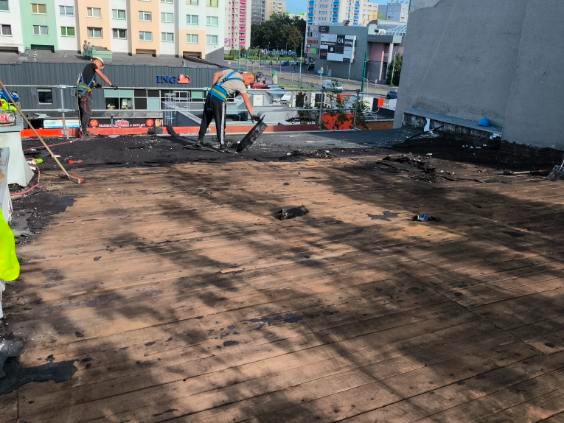 Pracownicy w uprzężach podczas prac na płaskim dachu budynku, prawdopodobnie związanych z jego uszczelnianiem lub naprawą. Widoczne narzędzia i materiały budowlane.