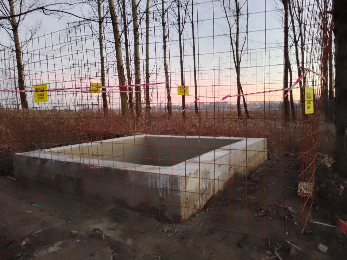 Betonowy zbiornik retencyjny w ziemi, otoczony siatką ochronną z tabliczkami ostrzegawczymi. Widoczne drzewa w tle, pochmurne niebo.