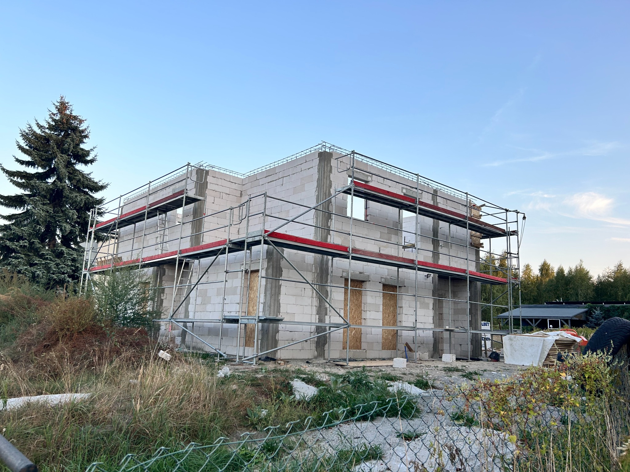 Surowy stan domu z białych bloczków, otoczonego rusztowaniem na tle błękitnego nieba i zieleni. Widoczne zabezpieczone okna i drzwi. Etap budowy.