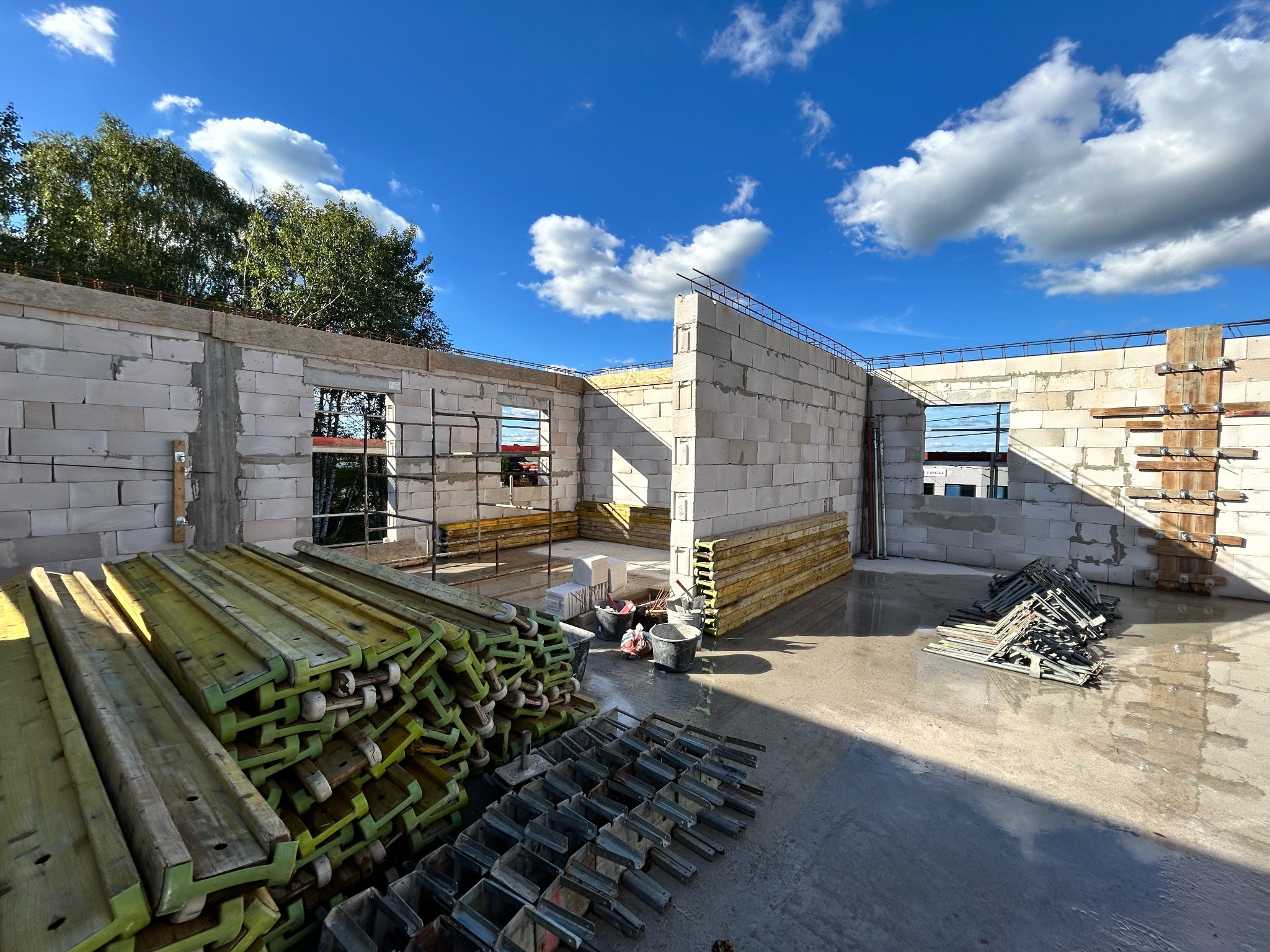 Wznoszenie ścian z betonu komórkowego Ytong. Widoczne szalunki, zbrojenia i narzędzia na mokrej posadzce. Budowa domu w słoneczny dzień.