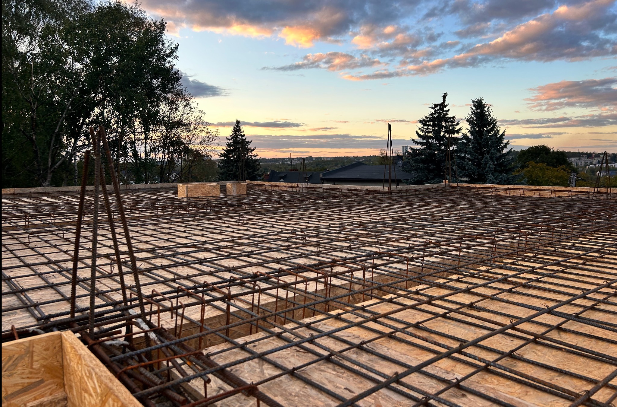Zbrojenie stropu na deskowaniu z widokiem na drzewa i zachodzące słońce. Stalowe pręty tworzą siatkę, przygotowane do zalania betonem. Budowa domu w trakcie realizacji.