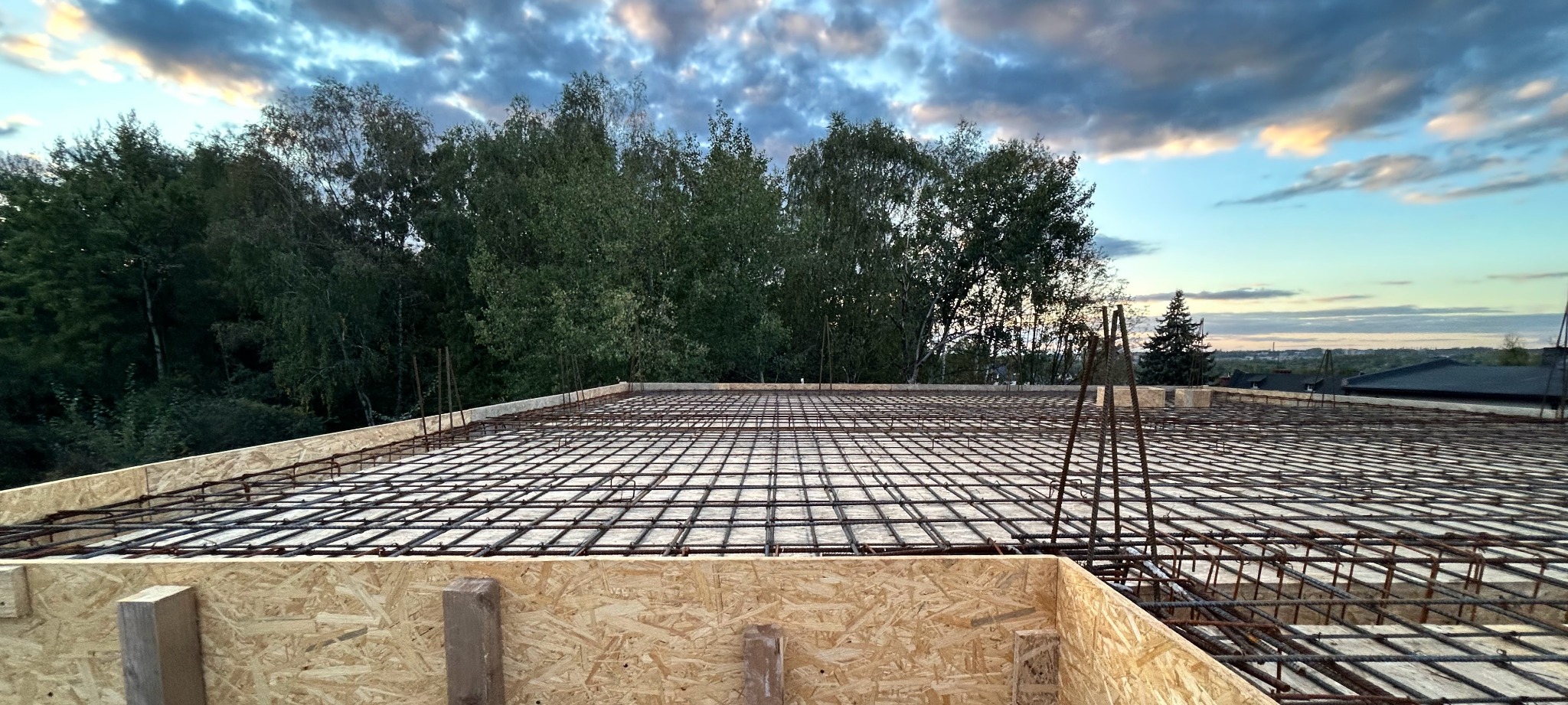 Zbrojenie stropu na drewnianym szalunku, widok z góry na siatkę z prętów stalowych. W tle drzewa i niebo o zmierzchu. Etap budowy domu.
