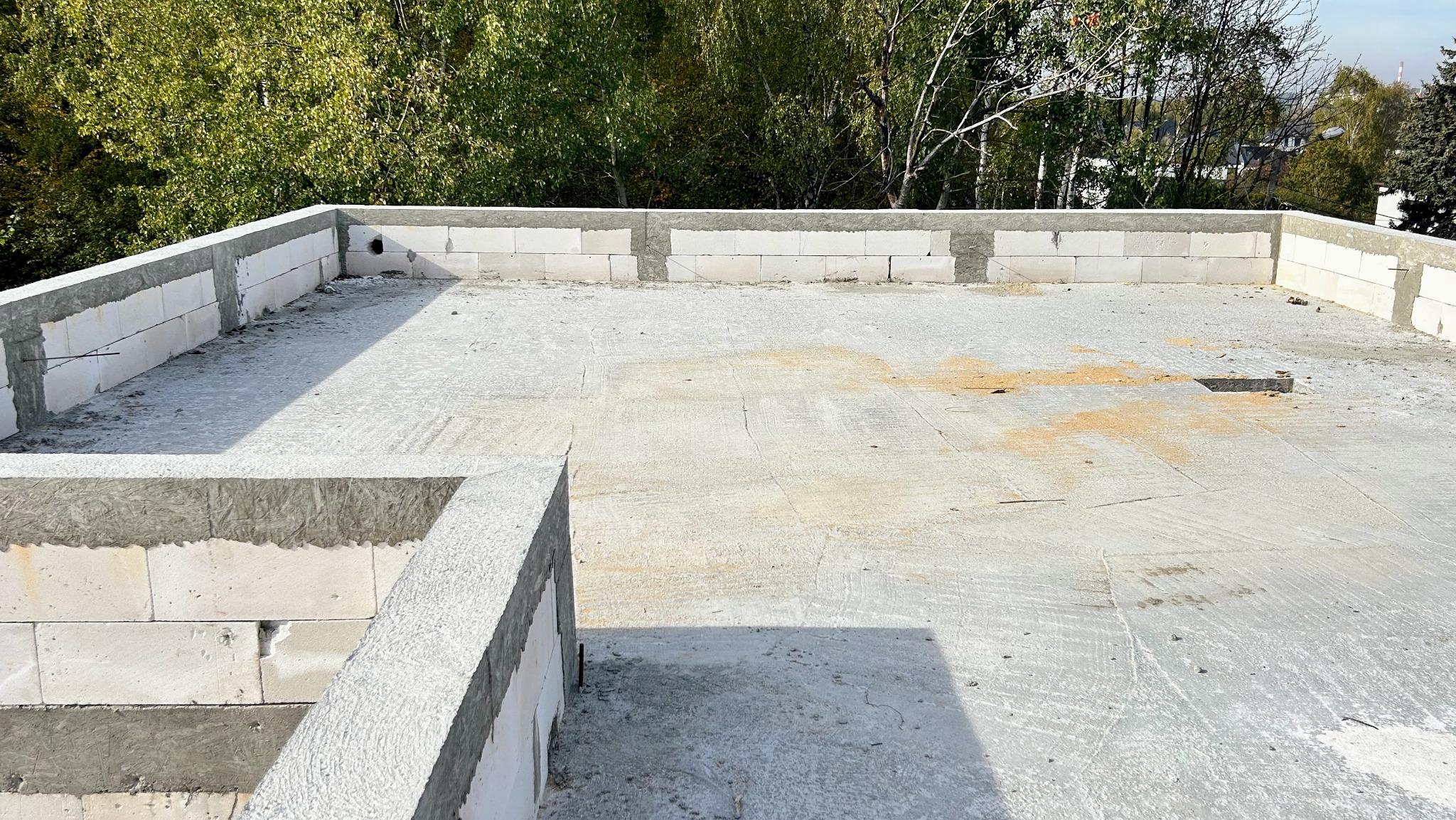 Surowy stan dachu budynku w trakcie budowy, z widocznymi bloczkami i betonową wylewką, na tle zieleni drzew. Widok z góry, w słoneczny dzień.