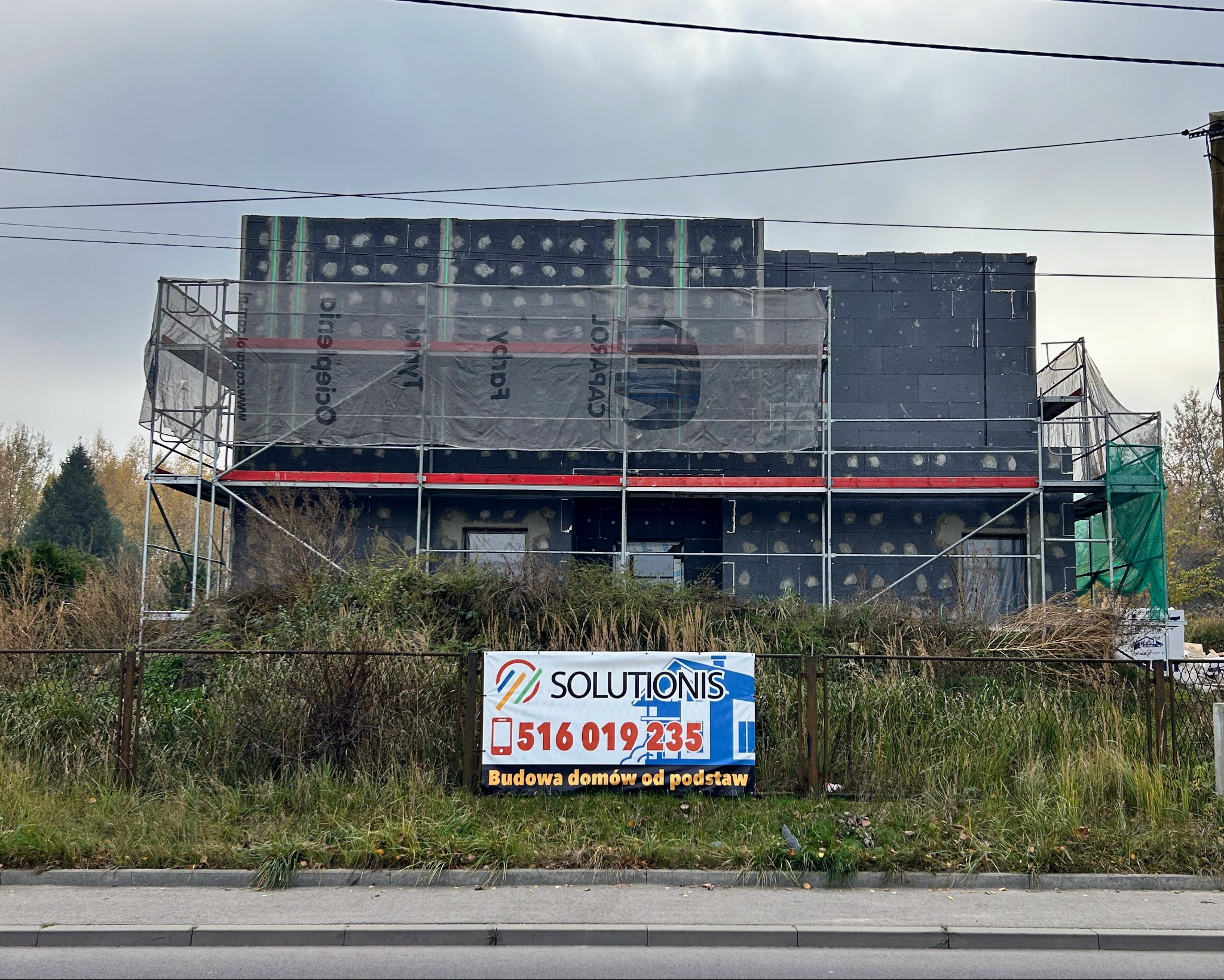 Budowa domu w trakcie ocieplania, widoczne rusztowania i czarne płyty izolacyjne. Na pierwszym planie baner reklamowy firmy budowlanej Solutionis.
