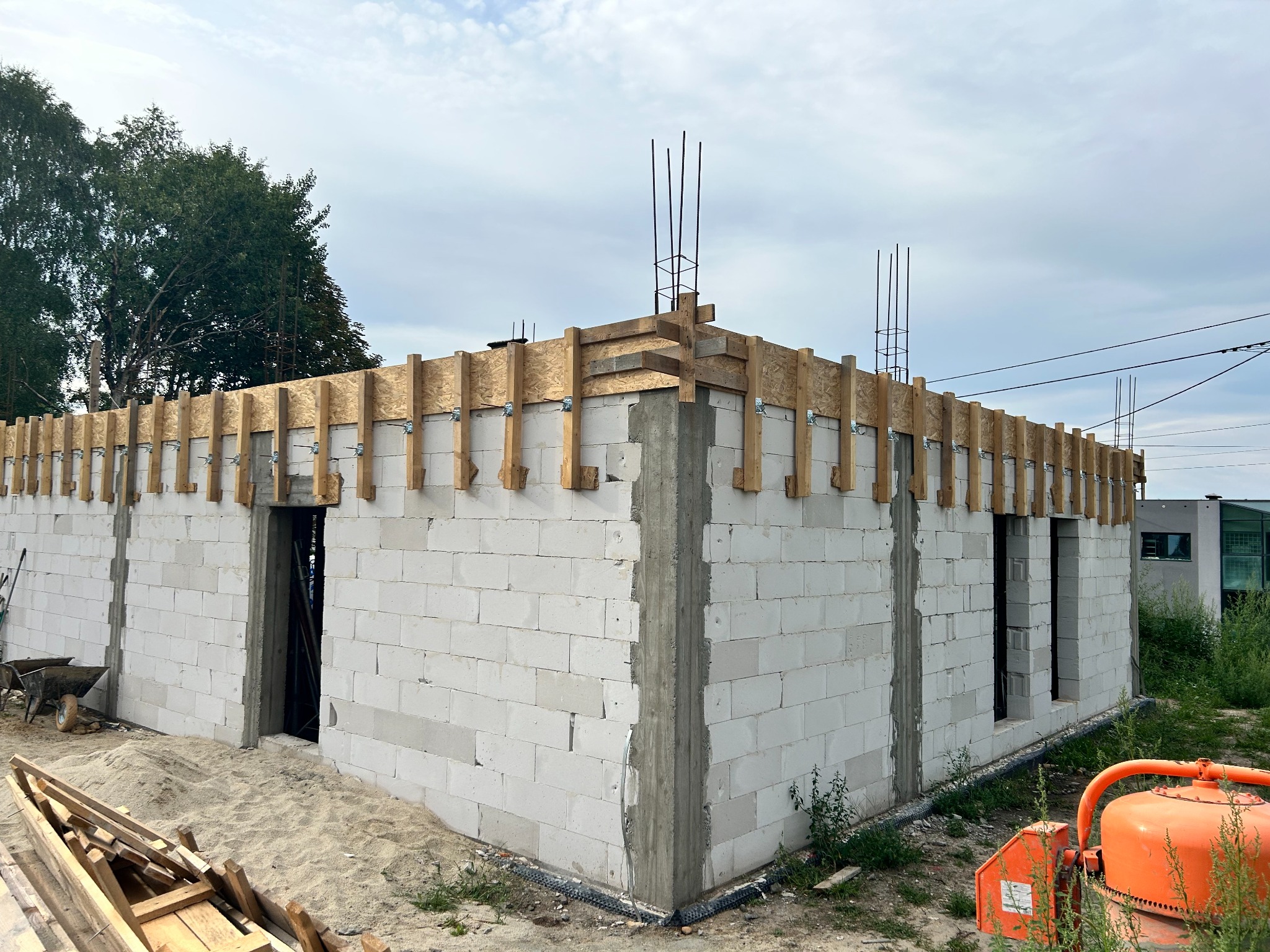 Budowa domu z białych bloczków, widoczne zbrojenia i szalunki. Na pierwszym planie pomarańczowa betoniarka, w tle drzewa i niebo.