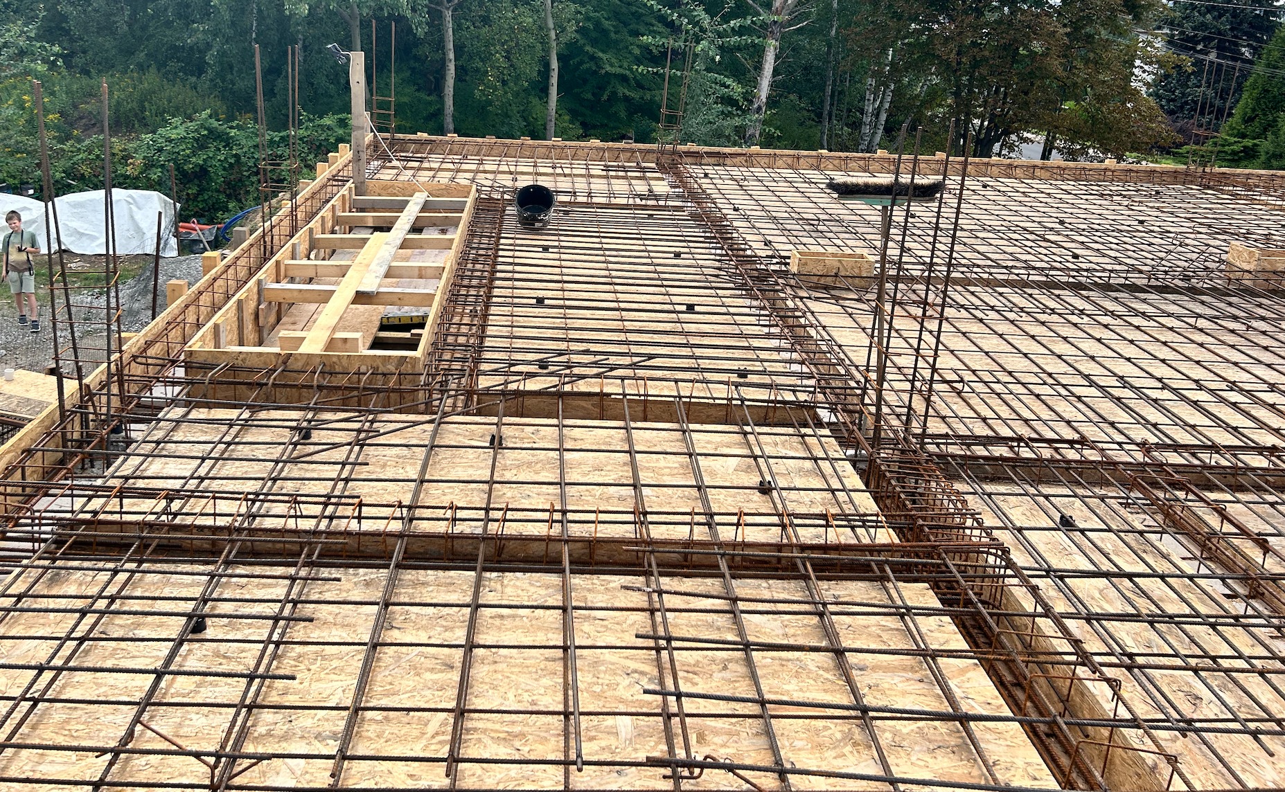 Zbrojenie stropu budynku w trakcie budowy. Widoczne drewniane szalunki i siatka z prętów stalowych. W tle zieleń drzew. Ujęcie z góry.
