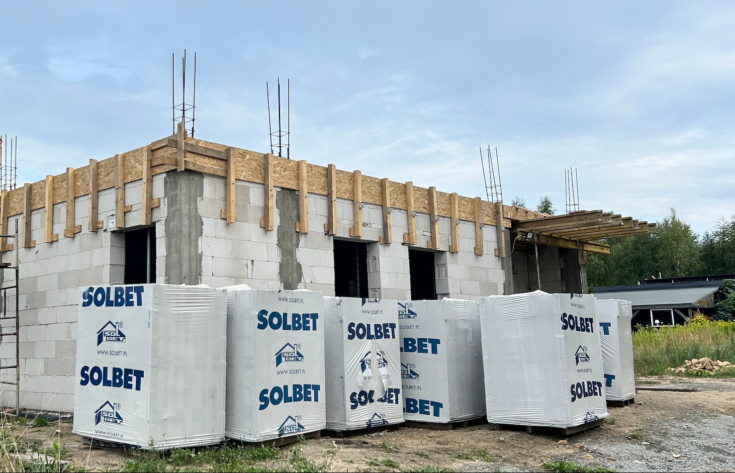 Budowa domu w stanie surowym otwartym z białych bloczków Solbet. Widoczne zbrojenia i drewniane szalunki. Na pierwszym planie palety z materiałem budowlanym.