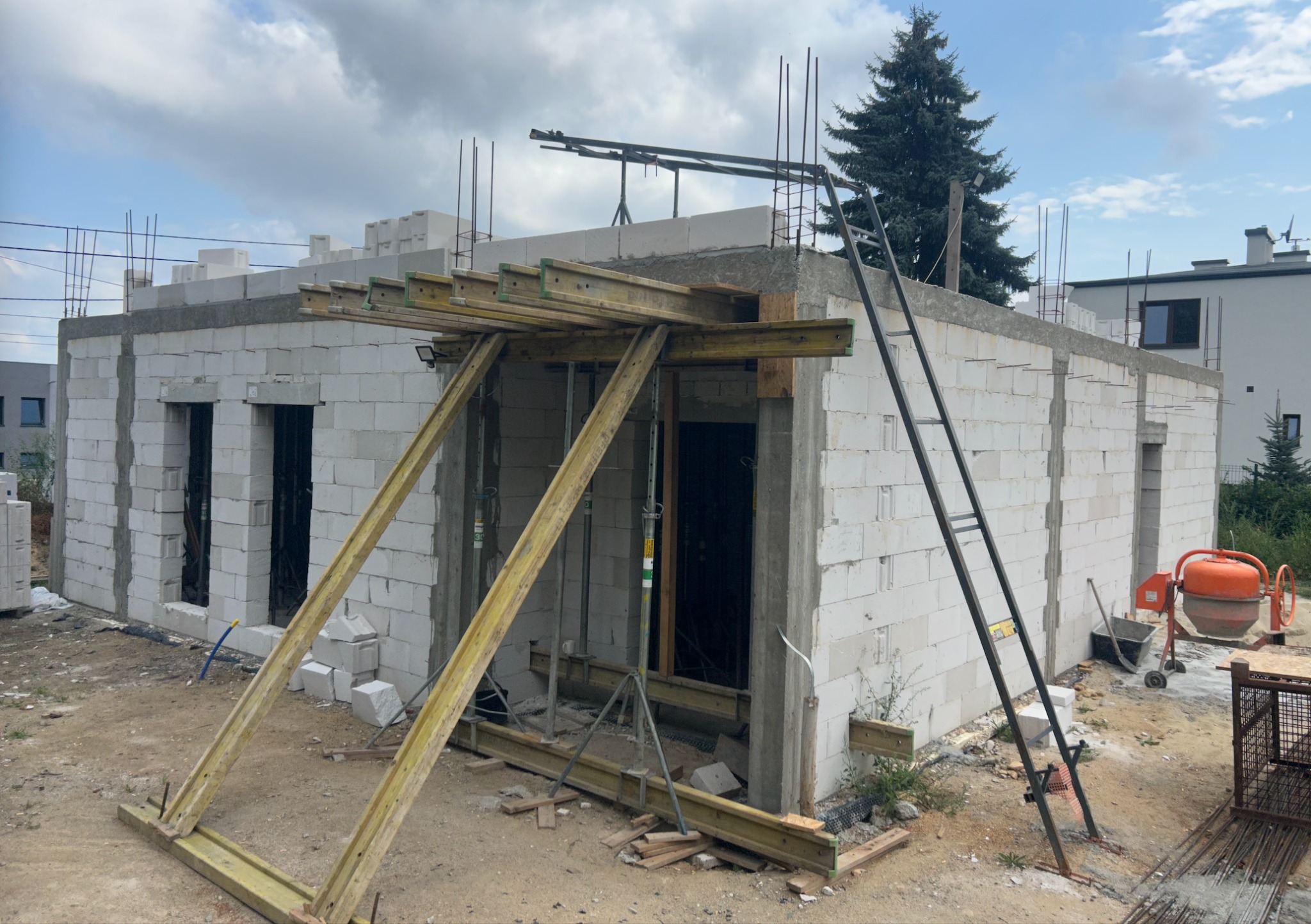 Budowa domu jednorodzinnego w trakcie realizacji: ściany z białych bloczków, szalunki drewniane, drabina oparta o ścianę, betoniarka w tle, widoczne zbrojenia.