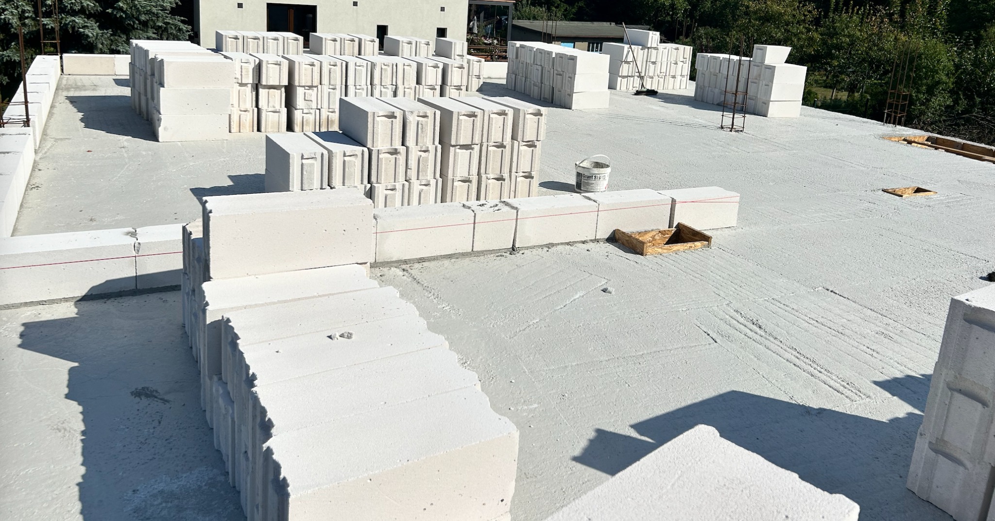 Wznoszenie ścian z białych bloczków na przygotowanym fundamencie. Widoczny fragment budynku w tle i wiadro z gruntem. Etap budowy domu.