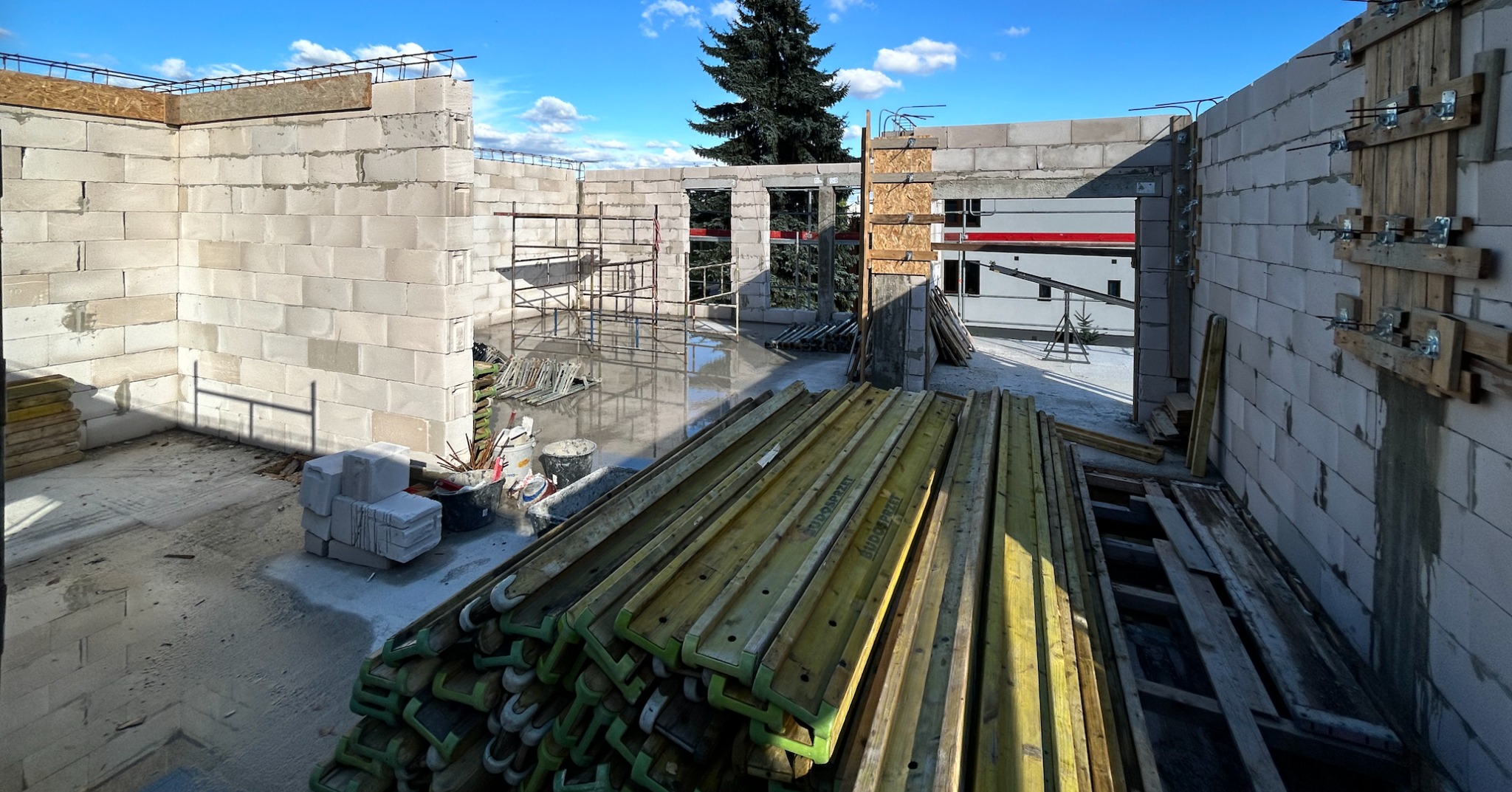 Wnętrze budynku w trakcie budowy z białych bloczków, widoczne rusztowania, szalunki i materiały budowlane. W tle zielone drzewo i błękitne niebo.