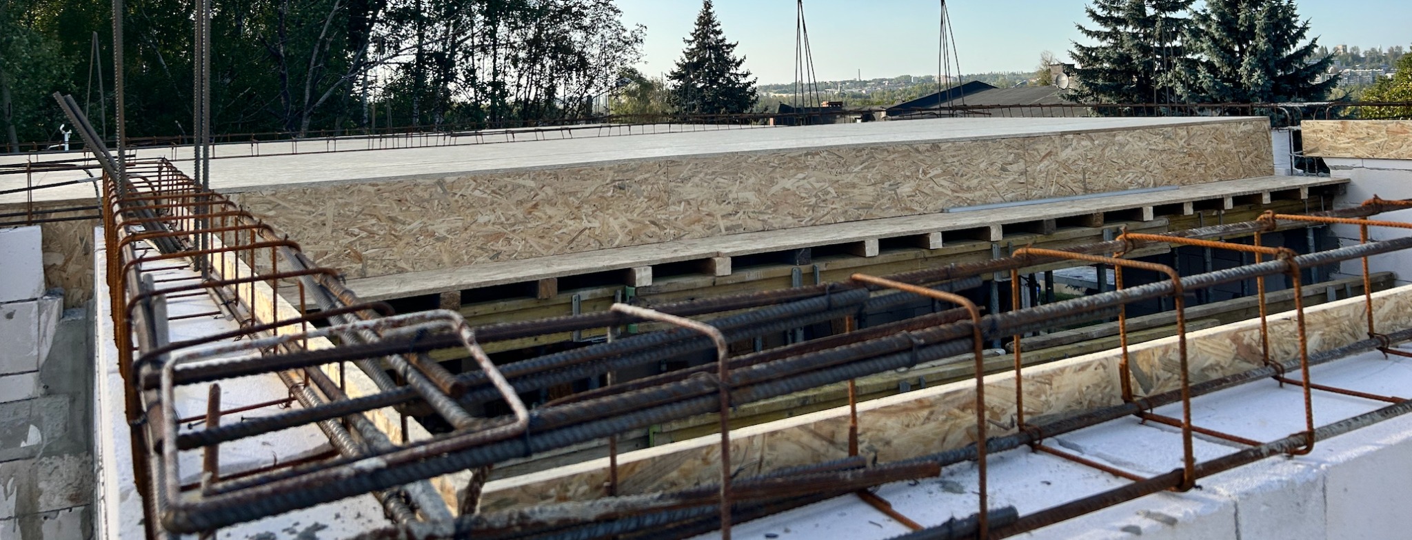 Zbrojenie stropu budynku w trakcie budowy, widoczne pręty zbrojeniowe i płyty OSB. W tle zieleń drzew i zabudowa miejska.