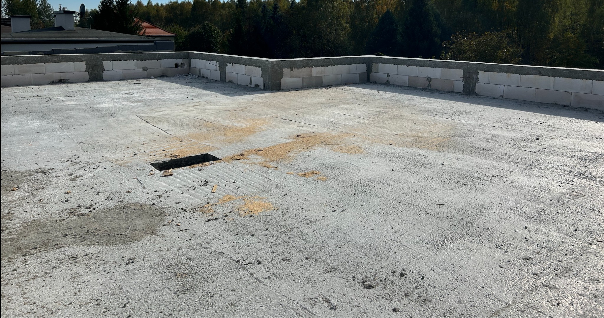 Surowy betonowy dach z fragmentem muru obwodowego z białych bloczków. Widoczny otwór w posadzce i ślady po budowie. W tle zieleń drzew i fragmenty budynków.