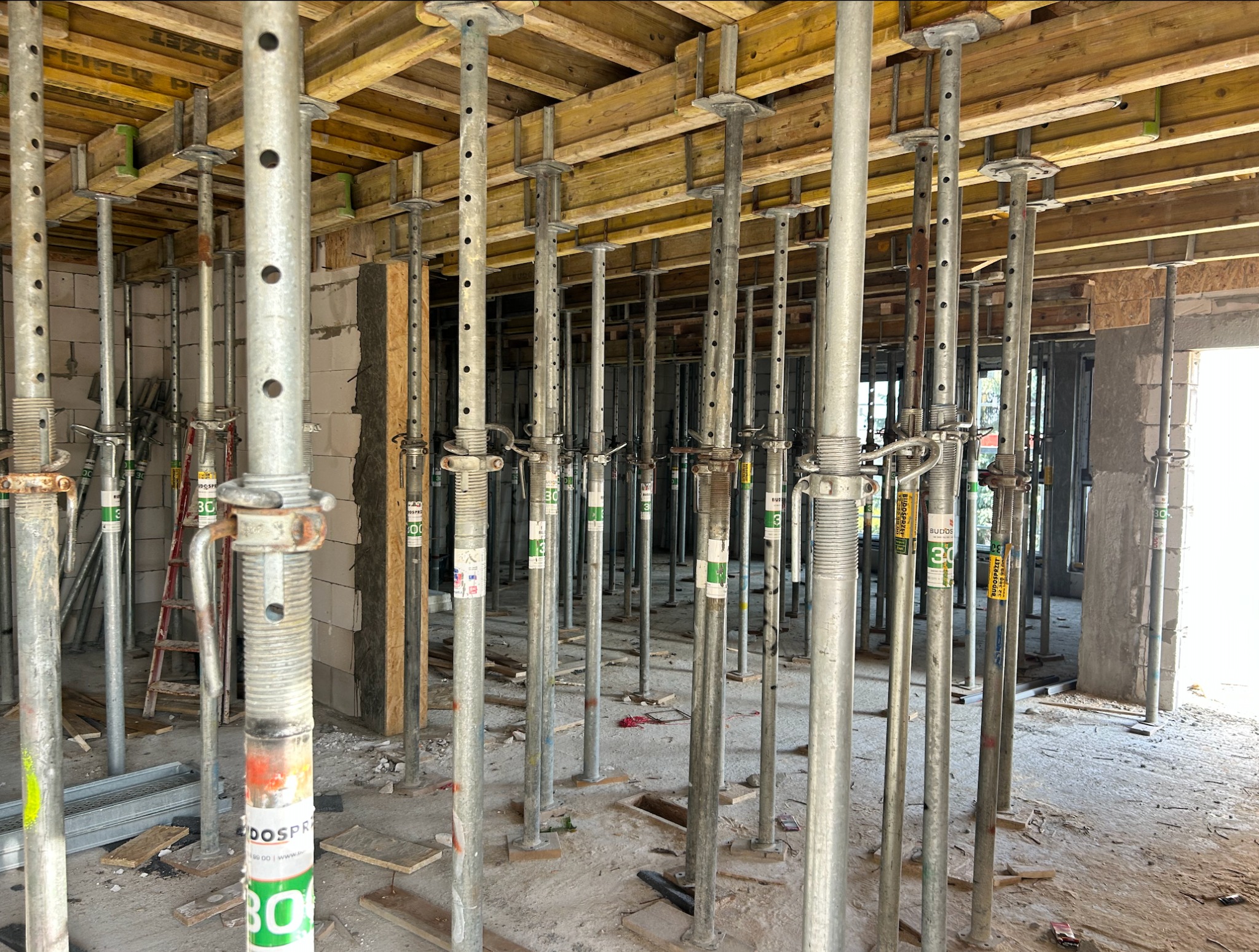 Wnętrze w trakcie budowy: liczne metalowe stemple podtrzymują drewniany strop, widoczne ściany z bloczków i drabina. Etap prac murarskich i konstrukcyjnych.