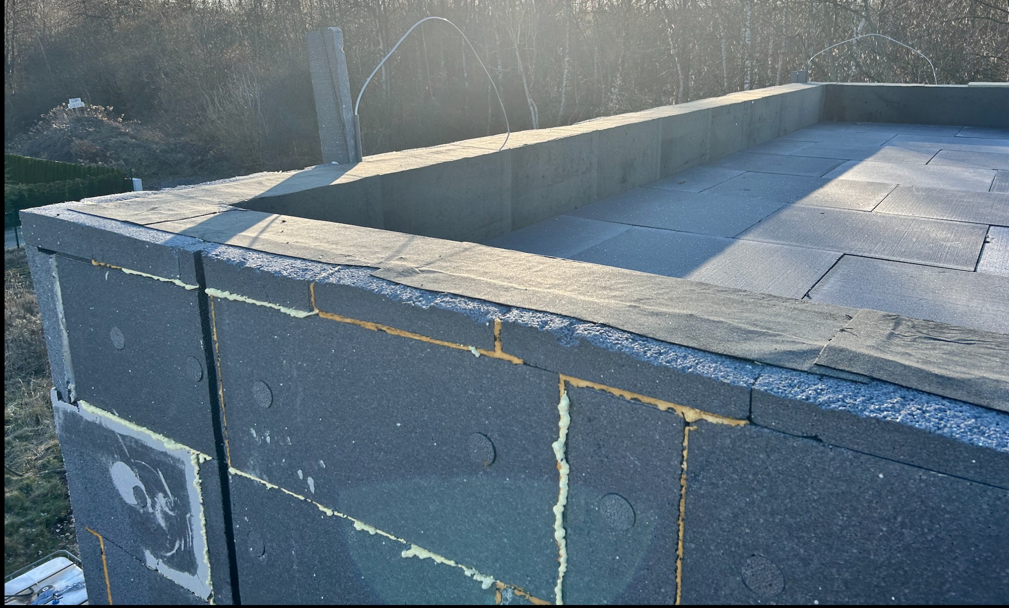 Ocieplenie budynku z szarego styropianu grafitowego, widoczne łączenia wypełnione pianką montażową. Na górze papa. W tle drzewa i niebo.