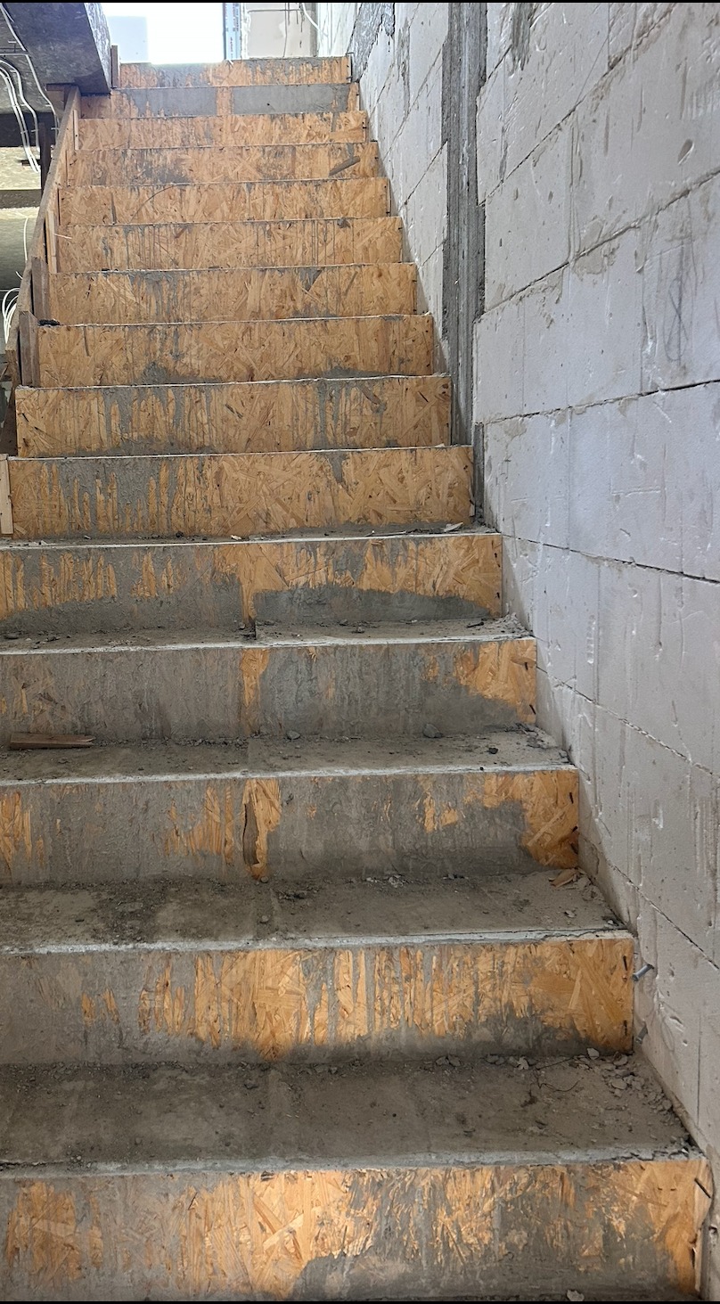 Surowe schody betonowe z drewnianym szalunkiem w trakcie budowy, widoczne bloczki betonowe na ścianie. Ujęcie z dołu, podkreślające pionowy charakter konstrukcji.