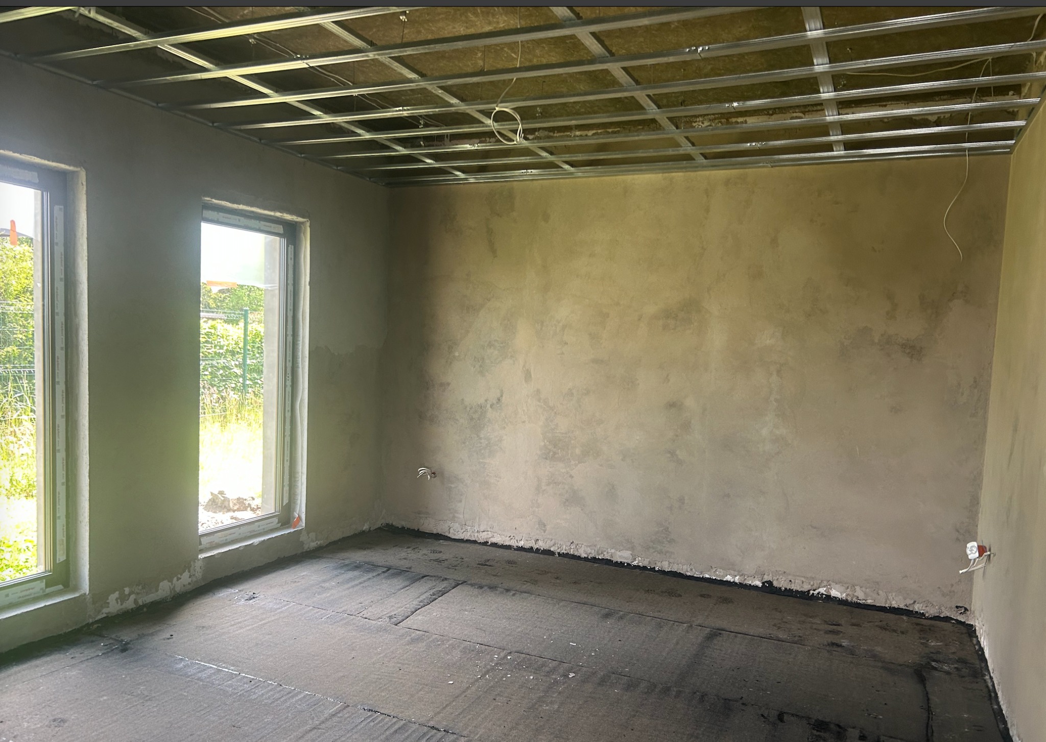 Surowy pokój w trakcie wykończenia: ściany po tynkowaniu, podłoga z izolacją, metalowa konstrukcja sufitu podwieszanego z widoczną wełną mineralną. Dwa okna z widokiem na zieleń.