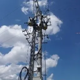 Słup energetyczny z zainstalowaną skrzynką elektryczną, widoczne przewody i elementy izolacyjne na tle błękitnego nieba z chmurami.