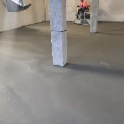 Świeżo wylana, szara posadzka w hali przemysłowej, z widocznymi słupami konstrukcyjnymi i pracownikiem wygładzającym powierzchnię za pomocą zacieraczki mechanicznej.