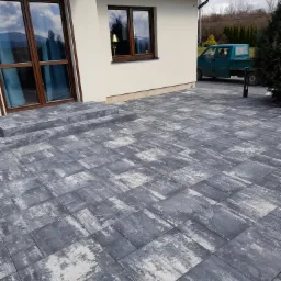 Ułożona kostka brukowa w odcieniach szarości i bieli prowadząca do schodów wejściowych domu z brązowymi oknami, na tle zielonego samochodu dostawczego.