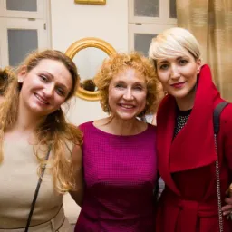 Trzy uśmiechnięte kobiety w różnym wieku, z blond włosami, w eleganckich ubraniach, pozują w jasnym wnętrzu z lustrem w złotej ramie w tle.