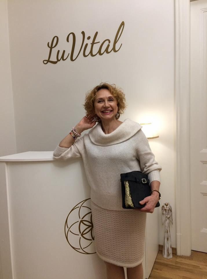 Kobieta z uśmiechem w salonie, opierająca się o białą ladę z logo, trzymająca czarną torebkę z złotymi elementami.