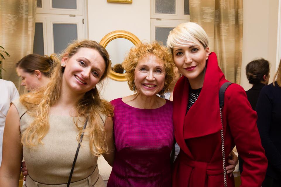 Trzy uśmiechnięte kobiety w różnym wieku, z blond włosami, w eleganckich ubraniach, pozują w jasnym wnętrzu z lustrem w złotej ramie w tle.