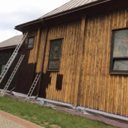 Fragment drewnianej elewacji budynku sakralnego, częściowo pomalowanej na brązowo, z trzema rozstawionymi drabinami i zabezpieczeniem folią malarską u podstawy, w tle widoczny fragment dachu...