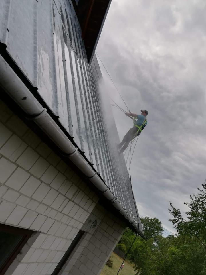 Pracownik zabezpieczony linami, myje pod ciśnieniem blaszany dach domu jednorodzinnego z białej cegły; widoczne okno i rynna.