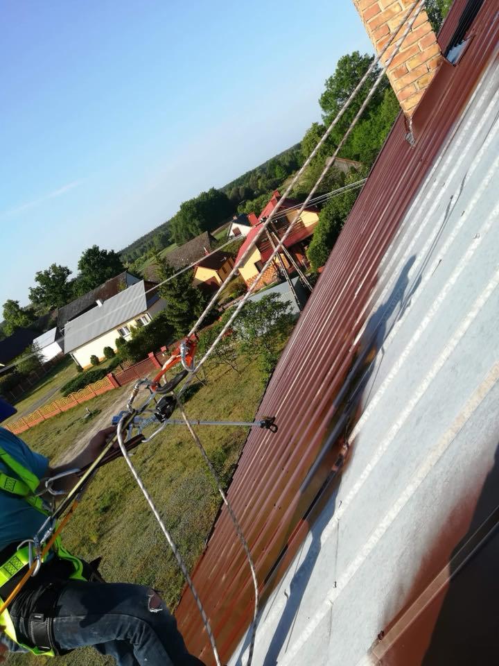 Pracownik zabezpieczony linami podczas pracy na dachu z blachy trapezowej, widoczne ślady czyszczenia, w tle zabudowania mieszkalne i drzewa.