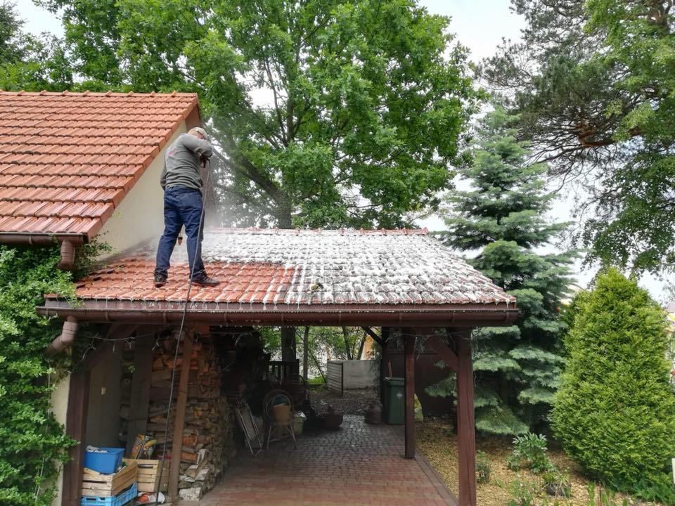 Mężczyzna w jeansach i szarej bluzie myje dach z czerwonej dachówki za pomocą myjki ciśnieniowej, widoczny efekt czyszczenia w postaci kontrastu między czystą a zabrudzoną częścią dachu.