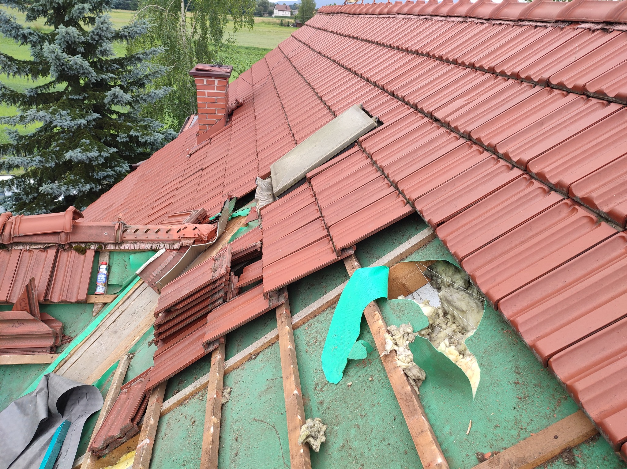 Częściowo zdjęte czerwone dachówki z odsłoniętą zieloną membraną i izolacją, widoczna konstrukcja drewniana, w tle komin z czerwonej cegły i drzewa
