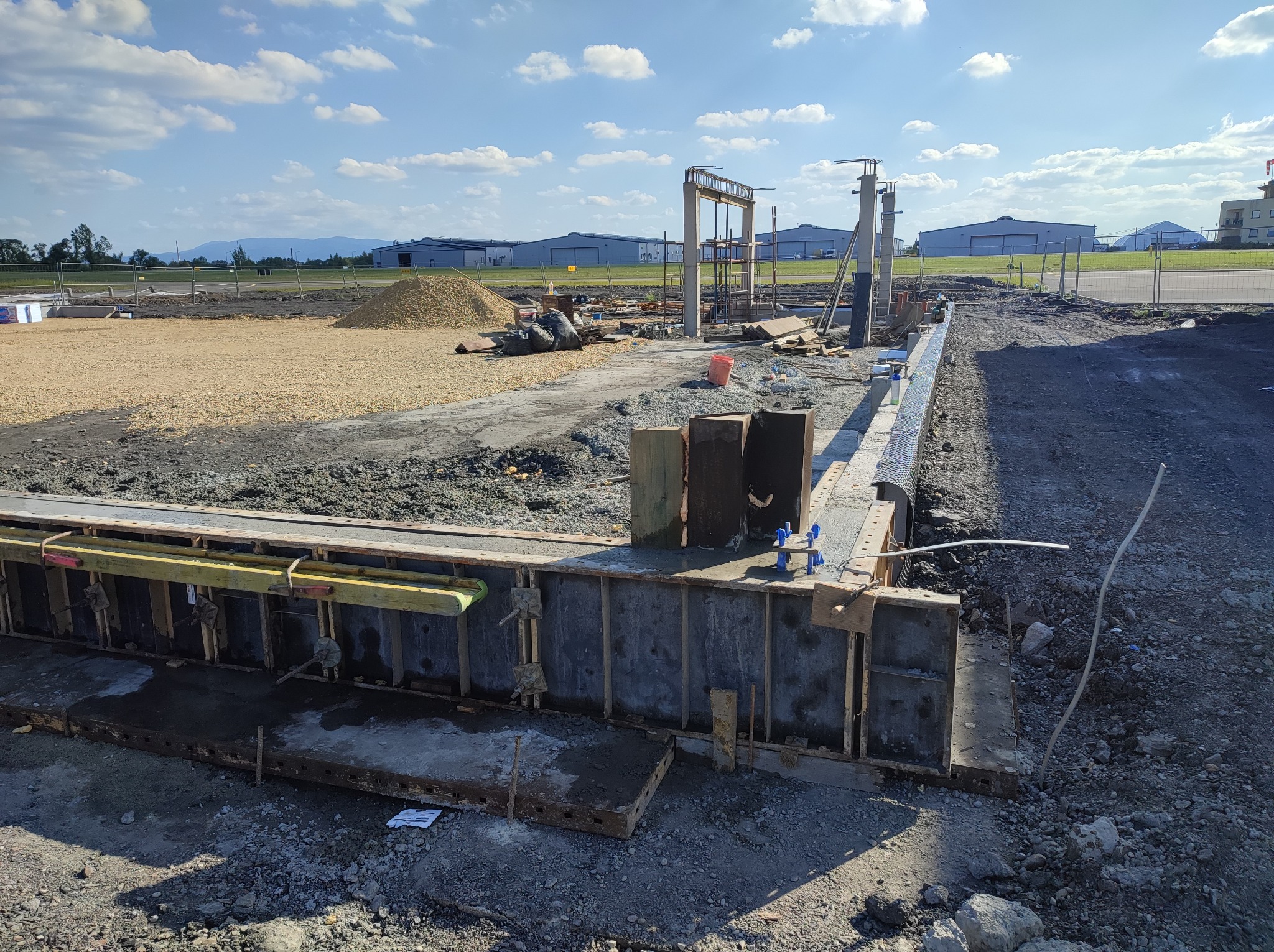 Szare, betonowe szalunki pod fundament z żółtymi elementami drewnianymi i metalowymi mocowaniami na placu budowy, w tle widoczne budynki lotniskowe i hałda żwiru.