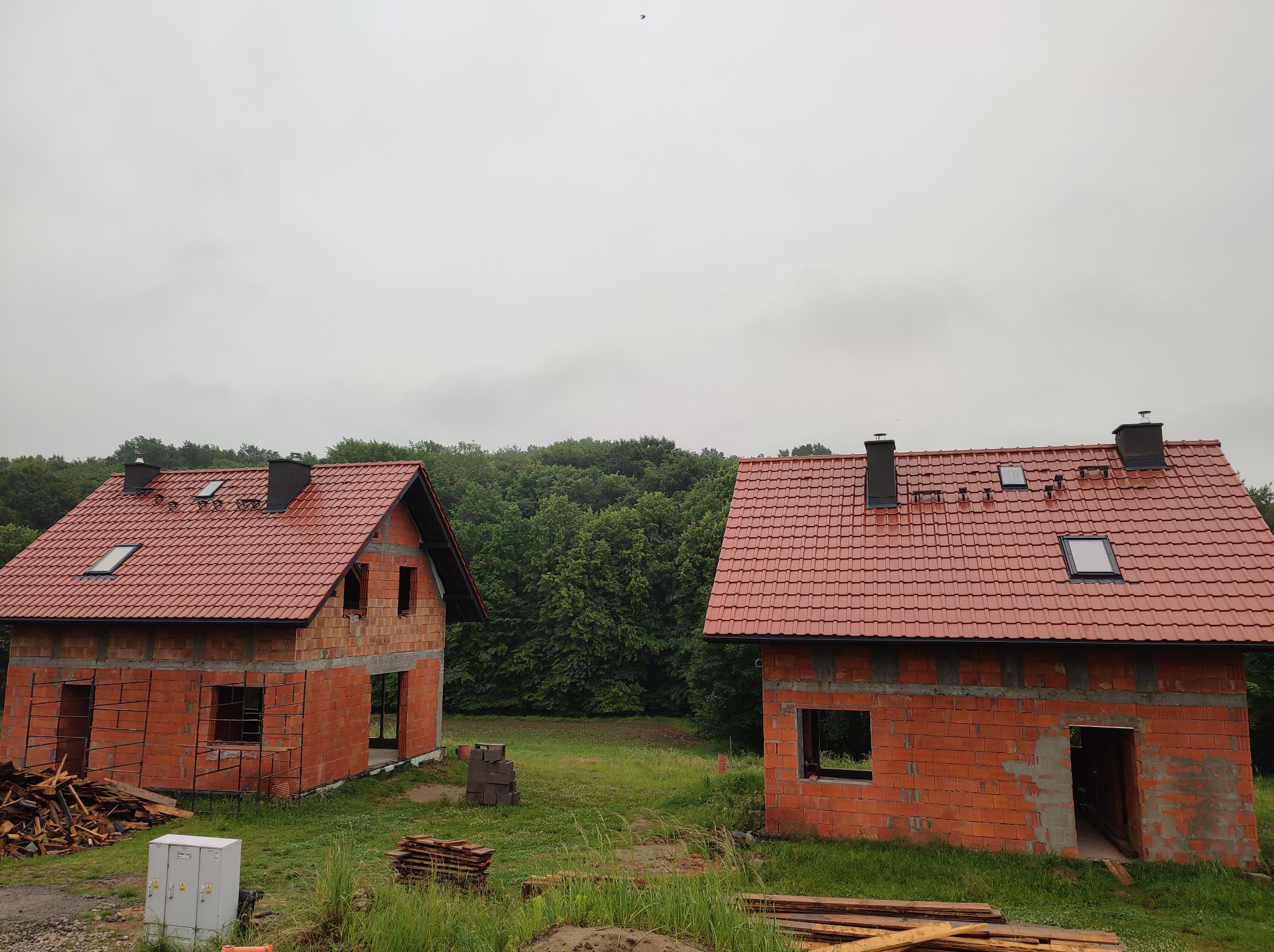 Dwa surowe, ceglane domy jednorodzinne z czerwoną dachówką w Krakowie, w trakcie budowy, z widoczną więźbą dachową i oknami dachowymi, na tle zieleni i pochmurnego nieba.