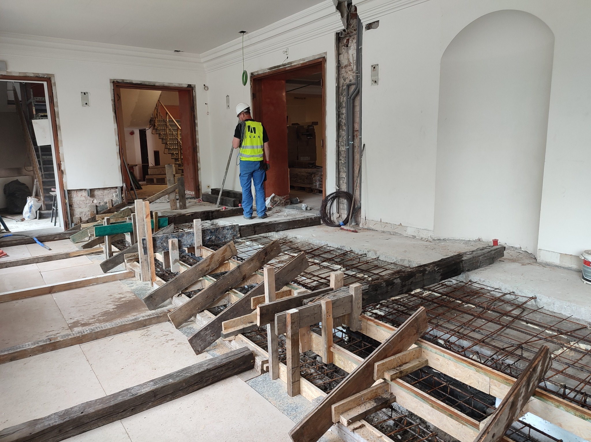 Przygotowanie zbrojenia i szalunku pod wylewkę fundamentu wewnątrz budynku, widoczny pracownik w kamizelce odblaskowej i kasku, zarysowane ściany i framugi drzwi.