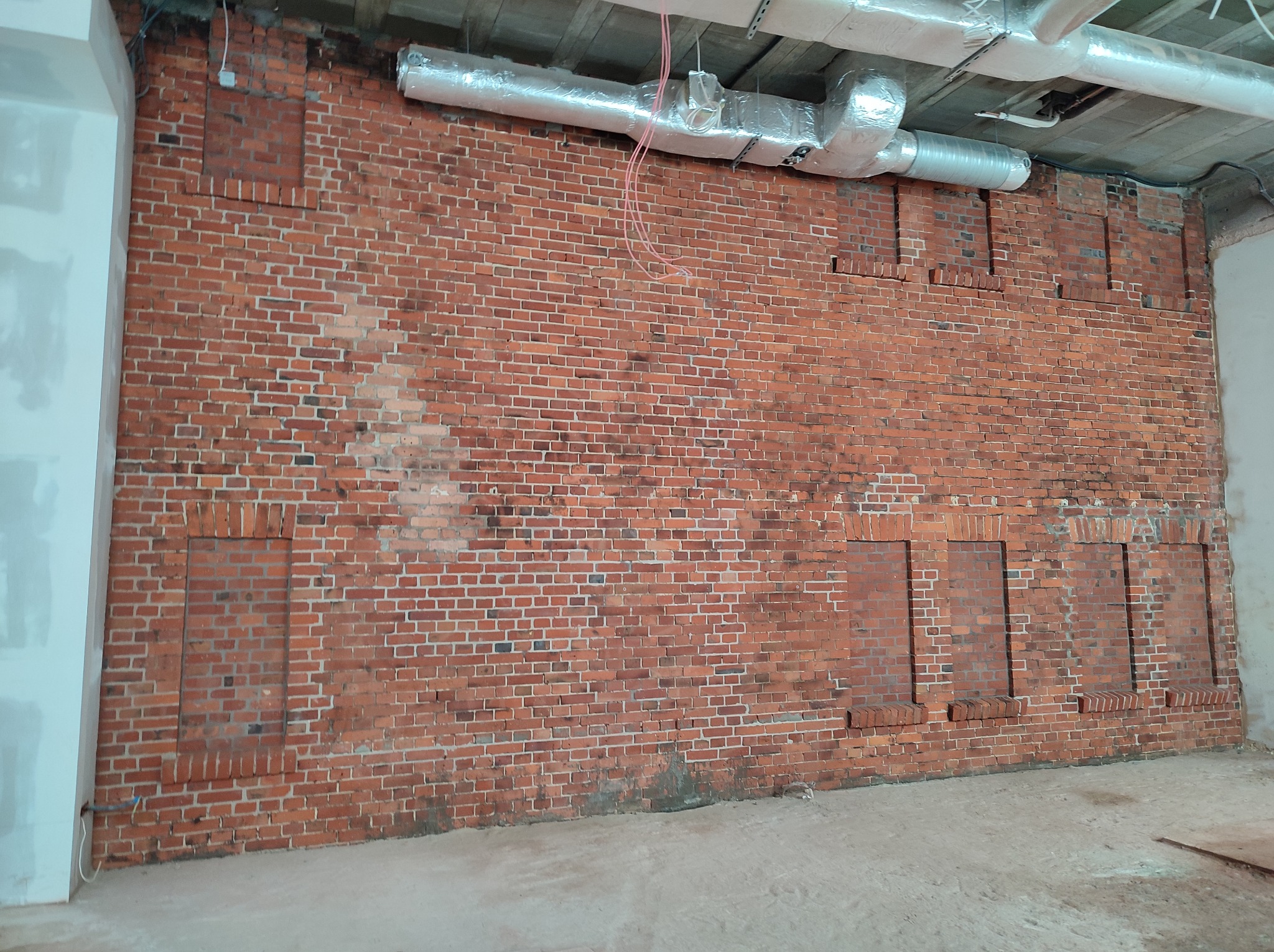 Czerwona ceglana ściana z widocznymi śladami po zamurowanych otworach, z instalacją wentylacyjną pod sufitem, w surowym stanie wykończenia.