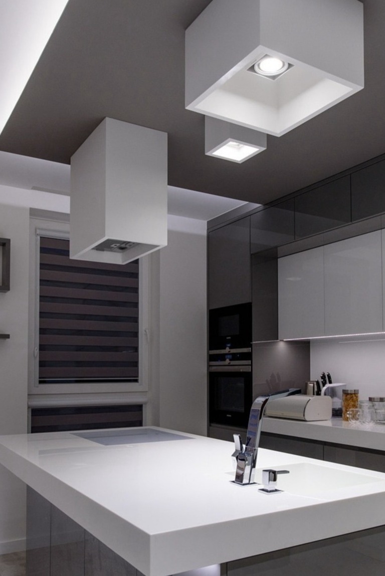Nowoczesna kuchnia z białym, minimalistycznym blatem wyspowym, chromowaną baterią, zabudową meblową w odcieniach szarości i oryginalnym oświetleniem sufitowym z kwadratowymi oprawami.