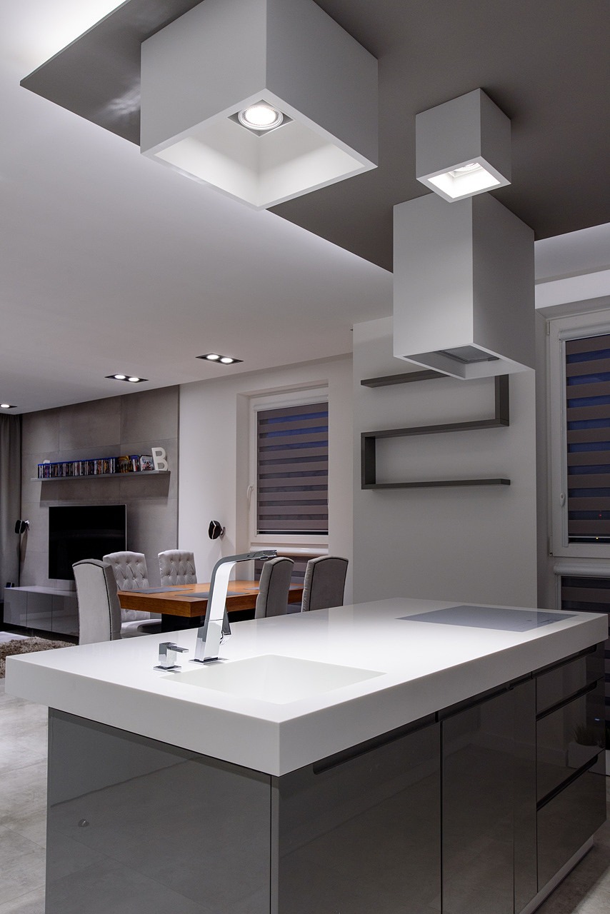 Nowoczesna kuchnia z wyspą, minimalistycznymi szafkami w kolorze szarym, białym blatem i designerską armaturą, oświetlona kwadratowymi lampami sufitowymi.