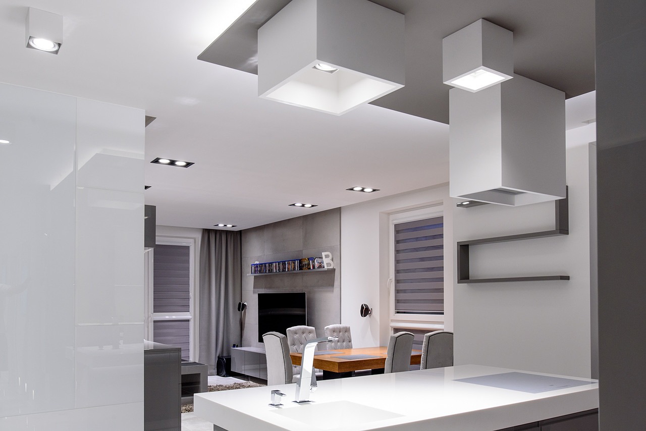 Nowoczesne wnętrze z otwartą przestrzenią: biała kuchnia z wyspą, drewniany stół jadalny otoczony szarymi krzesłami, szara ściana telewizyjna z półką, białe, kubiczne lampy sufitowe.