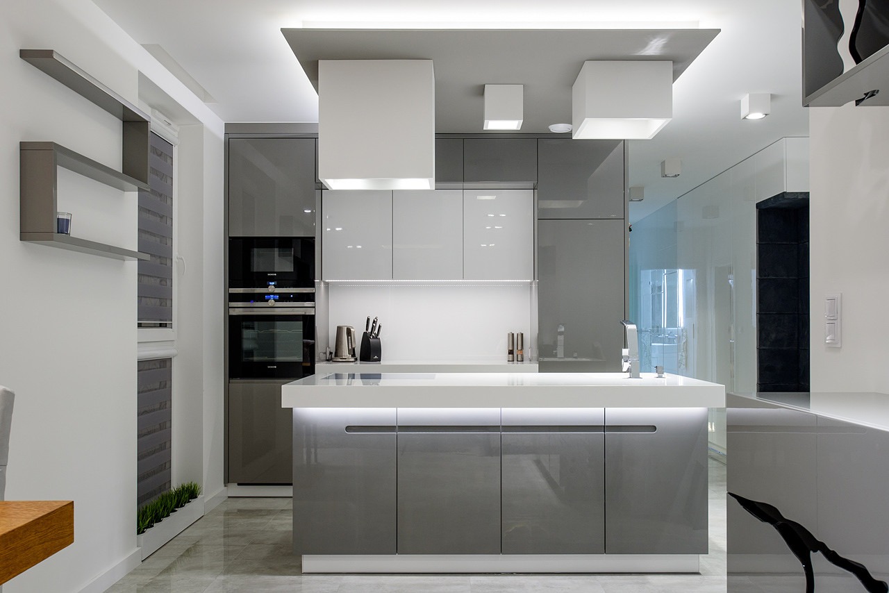 Nowoczesna kuchnia w odcieniach szarości i bieli z podświetlanym blatem, widoczne wbudowane sprzęty AGD, minimalistyczne oświetlenie sufitowe i dekoracyjne półki ścienne.