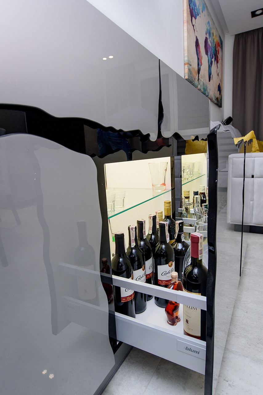 Nowoczesna szafka barowa z czarnymi, lakierowanymi frontami o nieregularnym kształcie, podświetlona półka z butelkami wina i szklankami, widoczny fragment salonu z obrazem na ścianie.