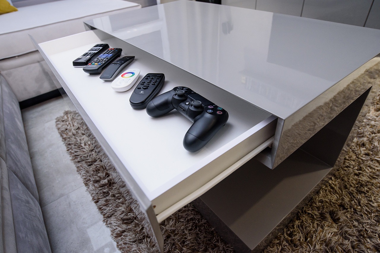 Nowoczesny stolik kawowy z wysuwaną szufladą, w której leżą piloty do urządzeń elektronicznych i kontroler do konsoli PlayStation.