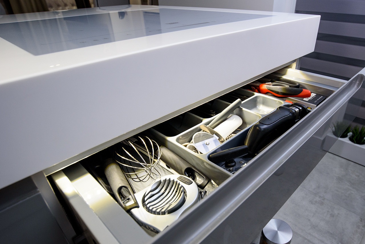 Otwarta szuflada w nowoczesnej kuchni z organizerem na przybory kuchenne, widoczna trzepaczka, nóż do pizzy i obieraczka.