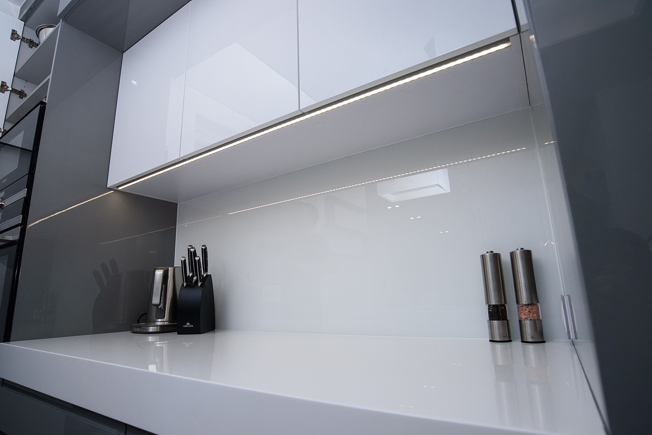 Nowoczesna kuchnia z białymi, błyszczącymi szafkami górnymi i blatem, podświetlona listwą LED, z zestawem noży w bloku i młynkami do soli i pieprzu na blacie.