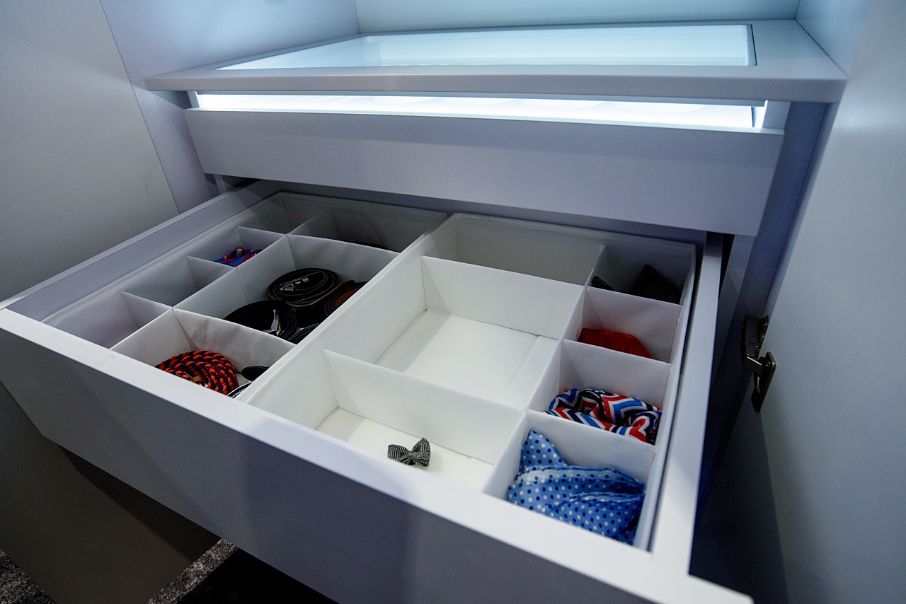 Otwarta biała szuflada z przegródkami, wypełniona kolorowymi akcesoriami męskimi, takimi jak krawaty, paski i muszki, podświetlana od góry.