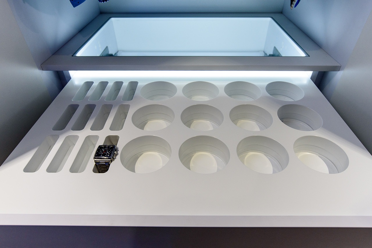 Biała, podświetlana szuflada z okrągłymi przegródkami i prostokątnymi wnękami, z jednym zegarkiem na bransolecie w jednej z wnęk.